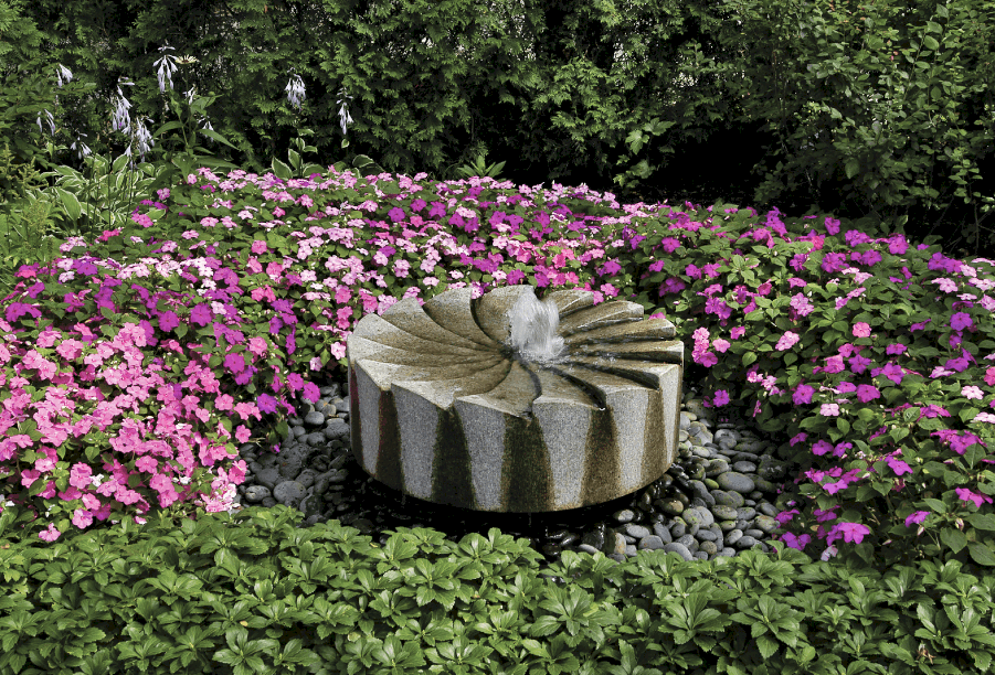 granite millstone fountain with Mexican blue pebbles and large bed of impatiens